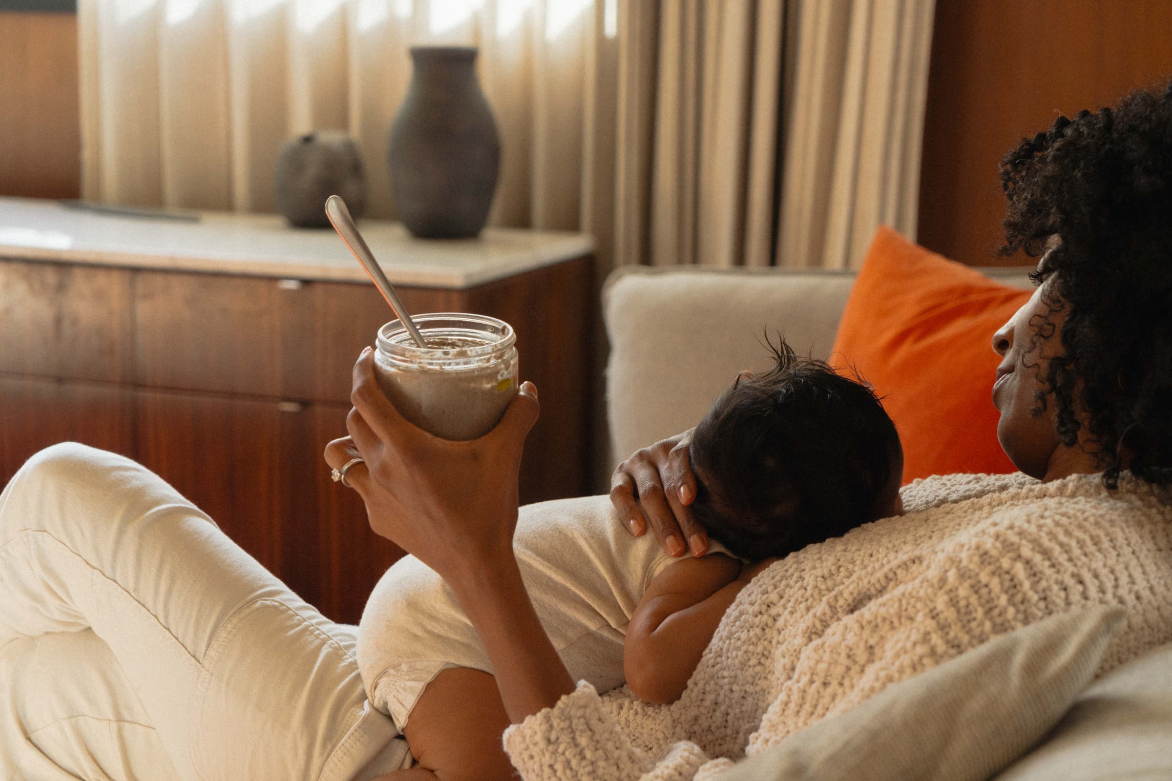 A mother resting on a sofa with her newborn, holding a Milky Oat jar